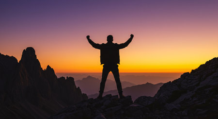 Silhouette of a hiker with raised hands on top of a mountain at sunsetの素材