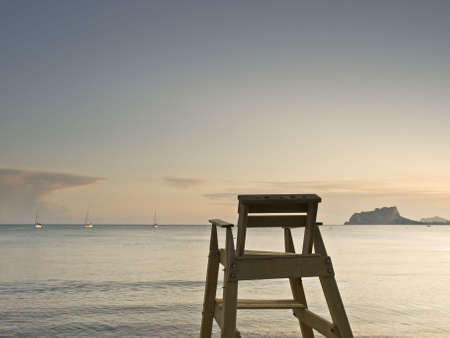 Seascape with a lifeguard chair on the foreground facing to the sea, with some sailboats and a mountain on the background の写真素材