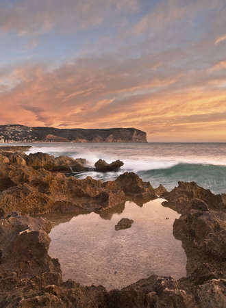 Seascape showing the sunrise on the Mediterranean coast, with a small pool of water on the rocky shore and a mountain in the background. Warm colours of sunrise in the sky.の写真素材