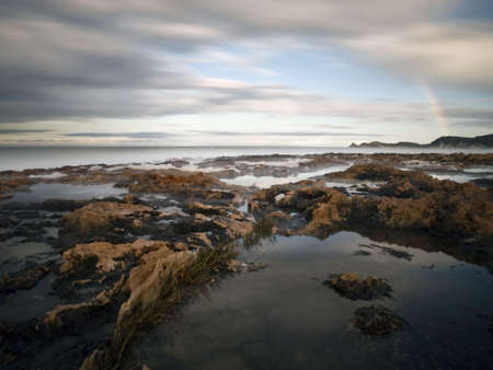 Mediterranean beach with a rainbow over the sea, with rocks on the foregroundの写真素材