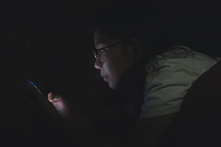 Asian woman using her mobile phone on the bed in dark room.の写真素材