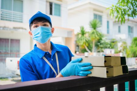 A smiling young asian delivery in blue uniform with parcel cardboard in front of customer house.の写真素材
