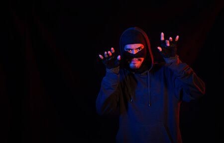 Robber, a thug in a balaclava raised his hands on a black background, surrenders. Robbery, hacker, crime, theft, arrest. Copy spaceの写真素材