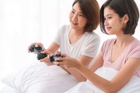 Two beautiful asian women who are best friends playing video game with wireless joystick on the bed in bedroom Both of her holidaysの写真素材