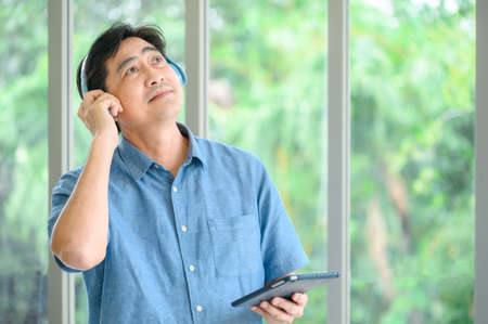 Senior male wearing head phones and lying back relaxing in an armchair with his eyes closed. He smiles and has one hand raised while being entertainedの写真素材