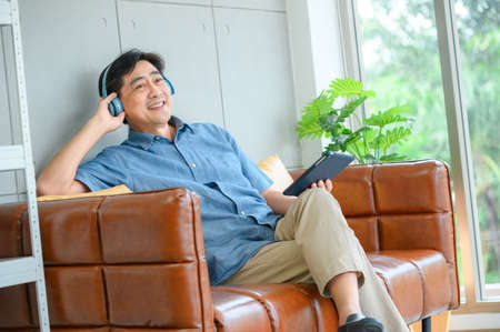 Senior male wearing head phones and lying back relaxing in an armchair with his eyes closed. He smiles and has one hand raised while being entertainedの写真素材