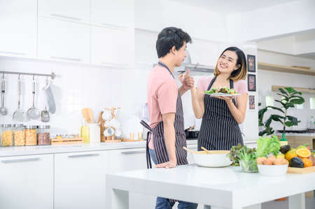 Cute joyful couple asian cooking together and adding spice to meal, laughing and spending time together in the kitchenの写真素材