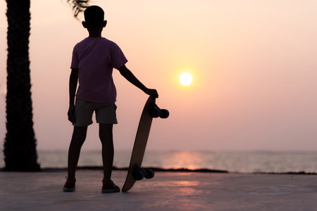 Boy standing with a surf skate, turned his back to the sunset.の写真素材