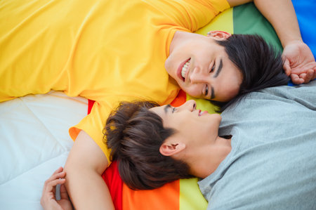 Male couple cuddling on bed in the morning. LGBQ Asian couple sleeping on colorful flags. Gender diversity symbolの写真素材