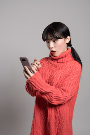 A studio portrait of a twenties Asian woman with a surprised look on the phoneの写真素材