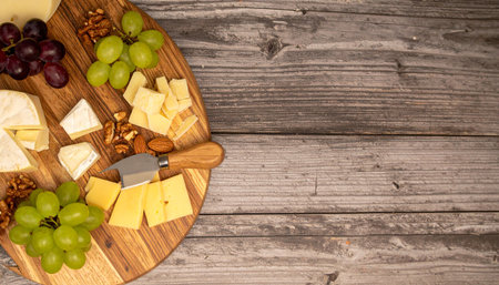 Variety of cheese, grapes, and nuts on a wooden platter, served on a rustic wooden table.の写真素材
