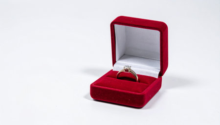 A diamond solitaire engagement ring sits in an open red velvet jewelry gift box on white background