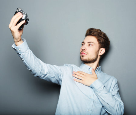 Handsome young man holding camera and making selfie and smiling while standing against grey...