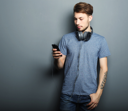 life style, happiness, emotional and people concept:  Handsome young man wearing headphones on his neck, holding mobile  and smiling while standing against grey backgroundの写真素材