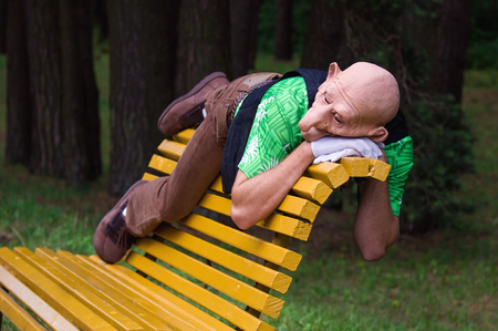 A man with a mask on a face lying on a yellow bench backrest in a park.の写真素材