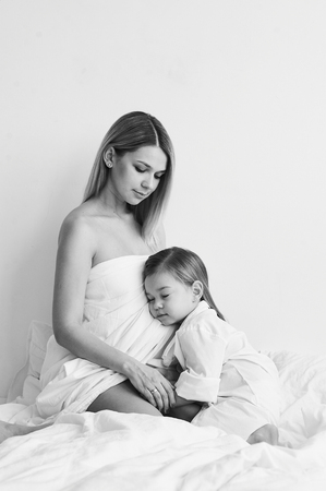 A beautiful pregnant mom and daughter playing on a white bed in the morning.の写真素材