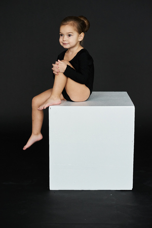 A portrait of beautiful little girl dressed in black tights on a white cube against dark background.の写真素材