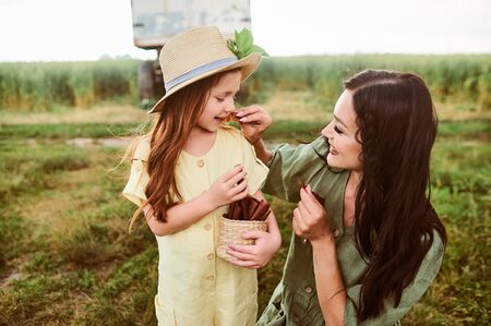 Beautiful young caucasian mother with her daughter in a linen dress with a basket of strawberries snacks gathers a new crop and has fun in the green fieldの写真素材