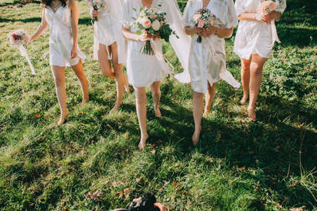 Faceless bridesmaids and bride in satin robes with bouquets in hands walk barefoot on the grass.の写真素材