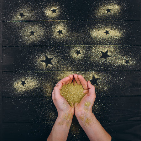 Human hands hold a shiny golden glitter in their palms and pour it forming stars on the table.