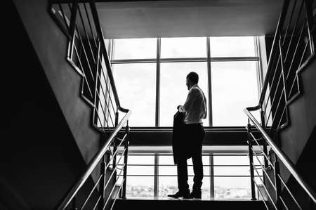 Black and white image of the groom looking out the window and holding a jacket on the stairs.の写真素材