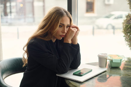 A young woman sits at a table in a cafÃ© with her eyes closed and hands on her temples, expressing...