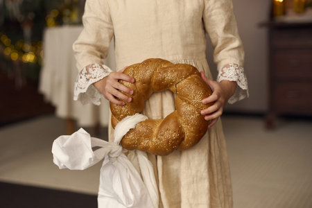 A young child dressed in a natural linen outfit holds a round, braided sesame bread wrapped with a...