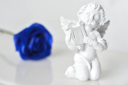 White ceramic figure of an angel on the white glossy surface and a blue rose behind in soft focus ,in the white background.の写真素材