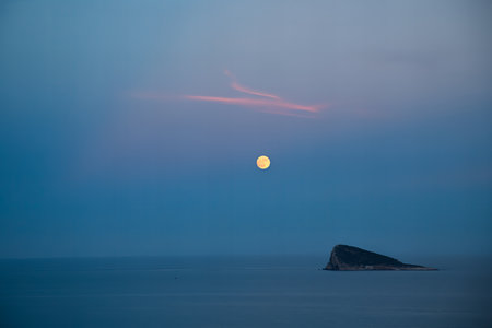 Serene full moon over calm ocean at dusk with small island in Benidorm, Spain.の写真素材