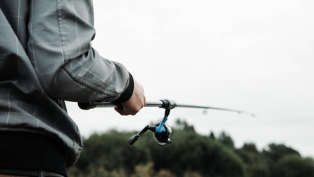Individual holding fishing rod, wearing gray jacket, standing by waters edge, surrounded by lush greenery, enjoying peaceful outdoor activity in natures serene environmentの写真素材