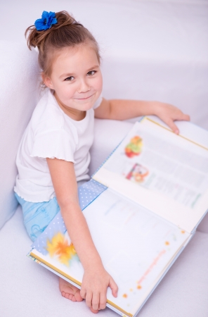 Cute little girl is reading book while sitting on a couch, indoor shootの写真素材