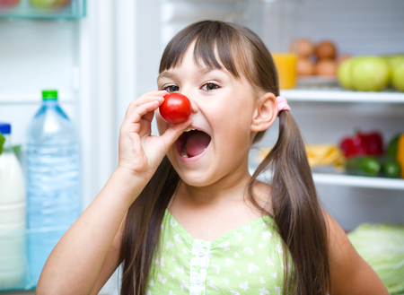 Happy girl eating tomatoes standing near refrigerator with fruits and vegetablesの写真素材