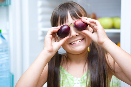 Happy girl eating plums standing near refrigerator with fruits and vegetablesの写真素材