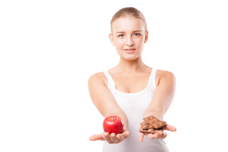 Doubtful woman holding an apple and chocolate trying to decide which one to eat on white backgroundの写真素材