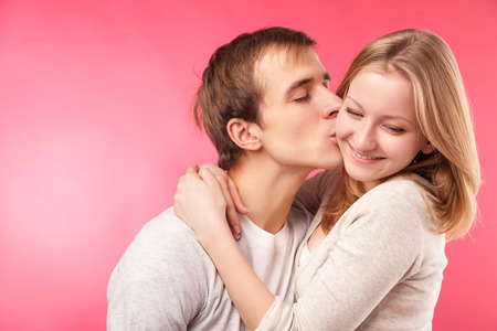 Close up studio portrait of young boy kissing cute girlfriend on cheek. Isolated on pink background.の写真素材