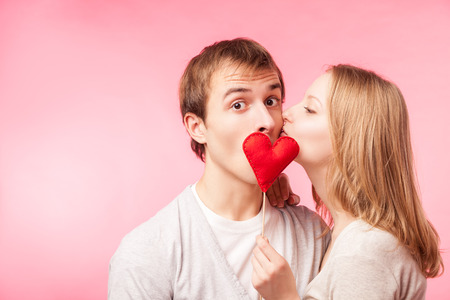 Young girl kissing boy hiding behind a little red heart. Isolated over pink background.の写真素材