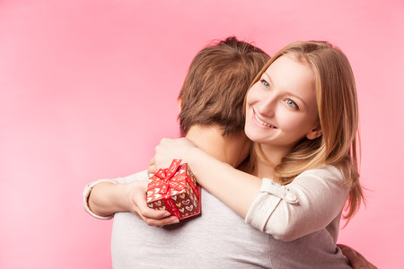 Young couple hugging with wrapped present. woman embracing man and holding present with ribbon. Isolated over pink background.の写真素材