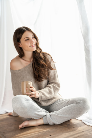 Beautiful girl with cup enjoying the freshness of the new day and looking through the window.の写真素材