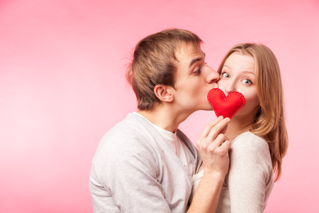 Young man kissing girl hiding behind a little red heart. Isolated over pink background.の写真素材