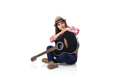 Young woman musician with guitar wearing shirt and hat sitting on a floor. White background.の写真素材