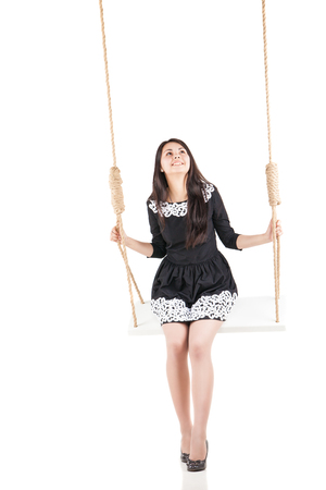 Beautiful girl in elegant black dress swinging on swing over white backgroundの写真素材