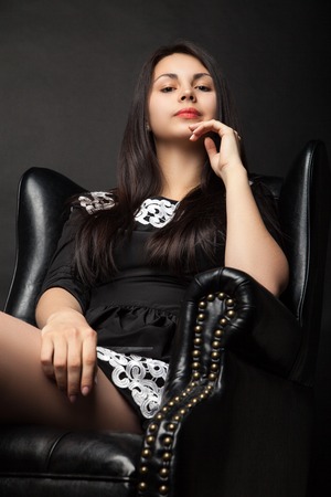 portrait of beautiful young woman in big black leather armchair over dark backgroundの写真素材