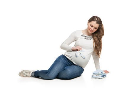 happy pregnant woman holding different items of child clothes over white backgroundの写真素材