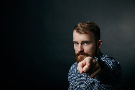 Red bearded handsome stylish man in blue checkered shirt, pointing, close up studio portrait on dark backgroundの写真素材