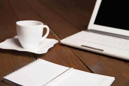 Close-up of ceramic cup with saucer,open notebook with laptop on wooden tableの写真素材