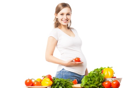 Pregnant smiling woman with tomato at table full of vegetables.Studio shotの写真素材