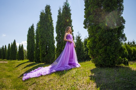 Blonde lady in violet dress with long skirt near row of green trees in sunlightの写真素材