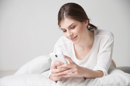 Smiling brunette using cell phone while lying on pillowの写真素材
