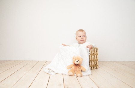 Smiling toddler sitting in bucket with blanket and toy bear looking away on white background.の写真素材