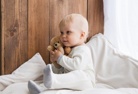 Sweet toddler looking away and hugging teddy bear sitting on white feather mattress near wooden wall.の写真素材
